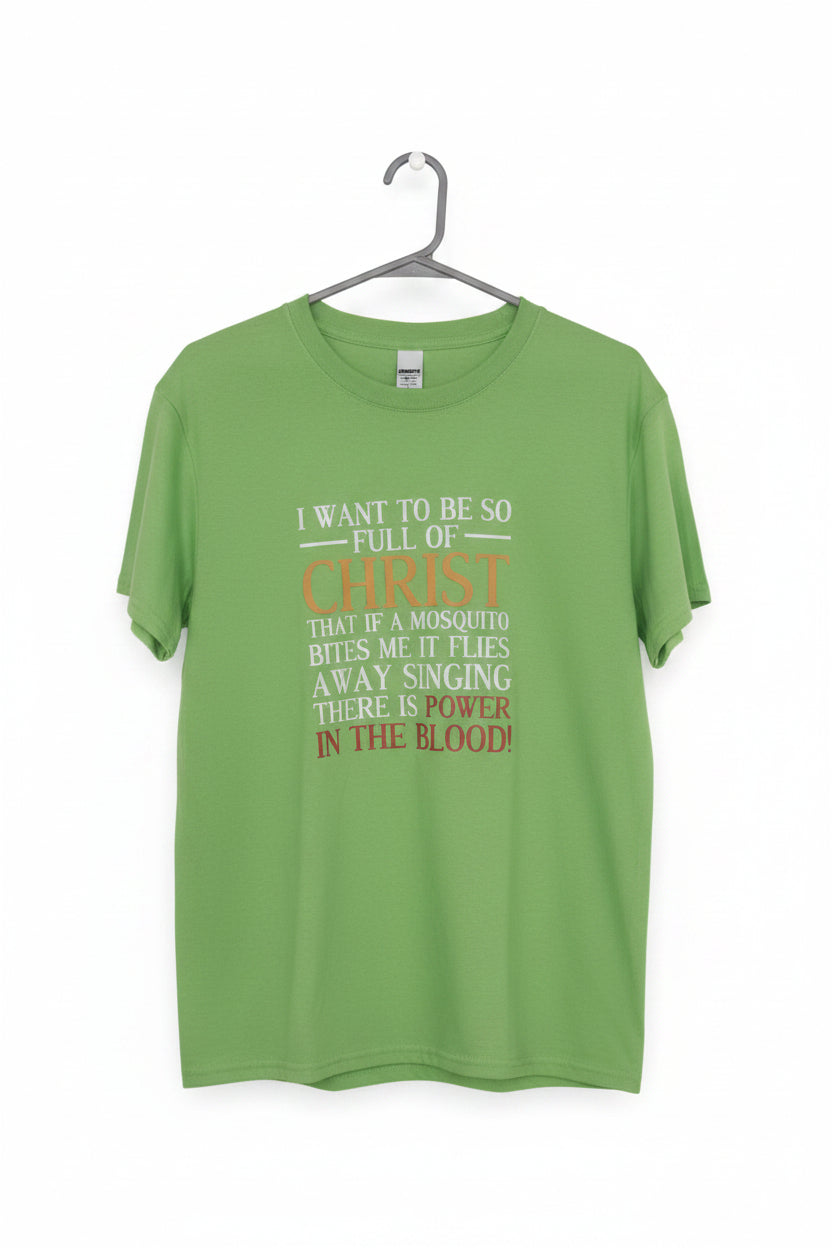 A bright green short-sleeve t-shirt hanging on a black metal hanger against a plain wall. The shirt features a humorous parody graphic with white and orange-red text that reads, 'I WANT TO BE SO FULL OF CHRIST THAT IF A MOSQUITO BITS ME IT FLYS AWAY SINGING THERE IS NO POWER IN THE BLOOD!'.